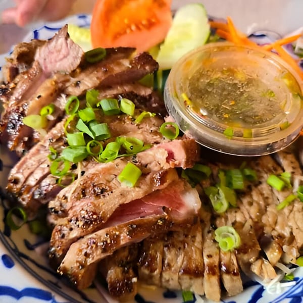 Sliced grilled beef with green onions, a side of vegetables, and dipping sauce on a patterned plate.